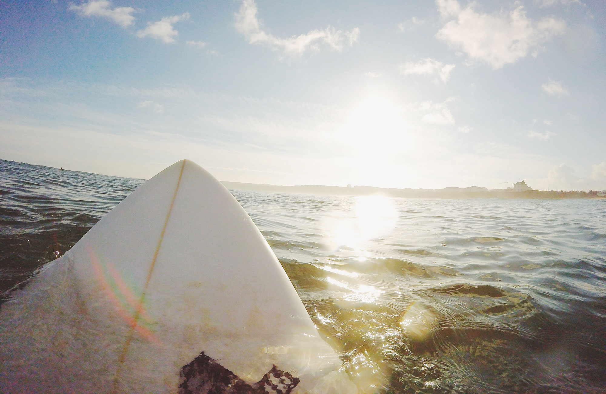 Image of the tip of a surfboard poking out of the water with the glare of the sun - KILROY