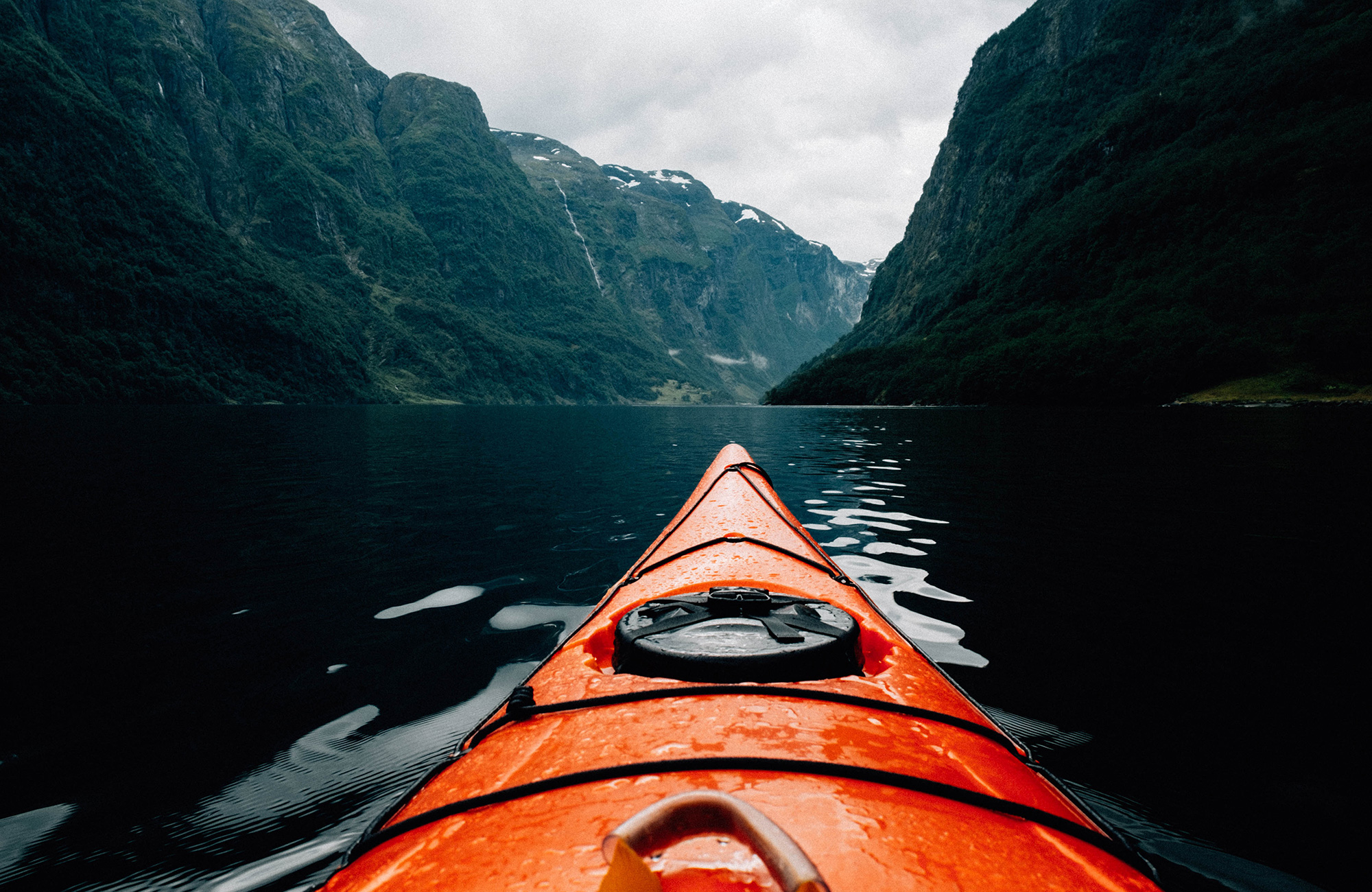 Image of the front of an orange kayak on the water between mountains - KILROY