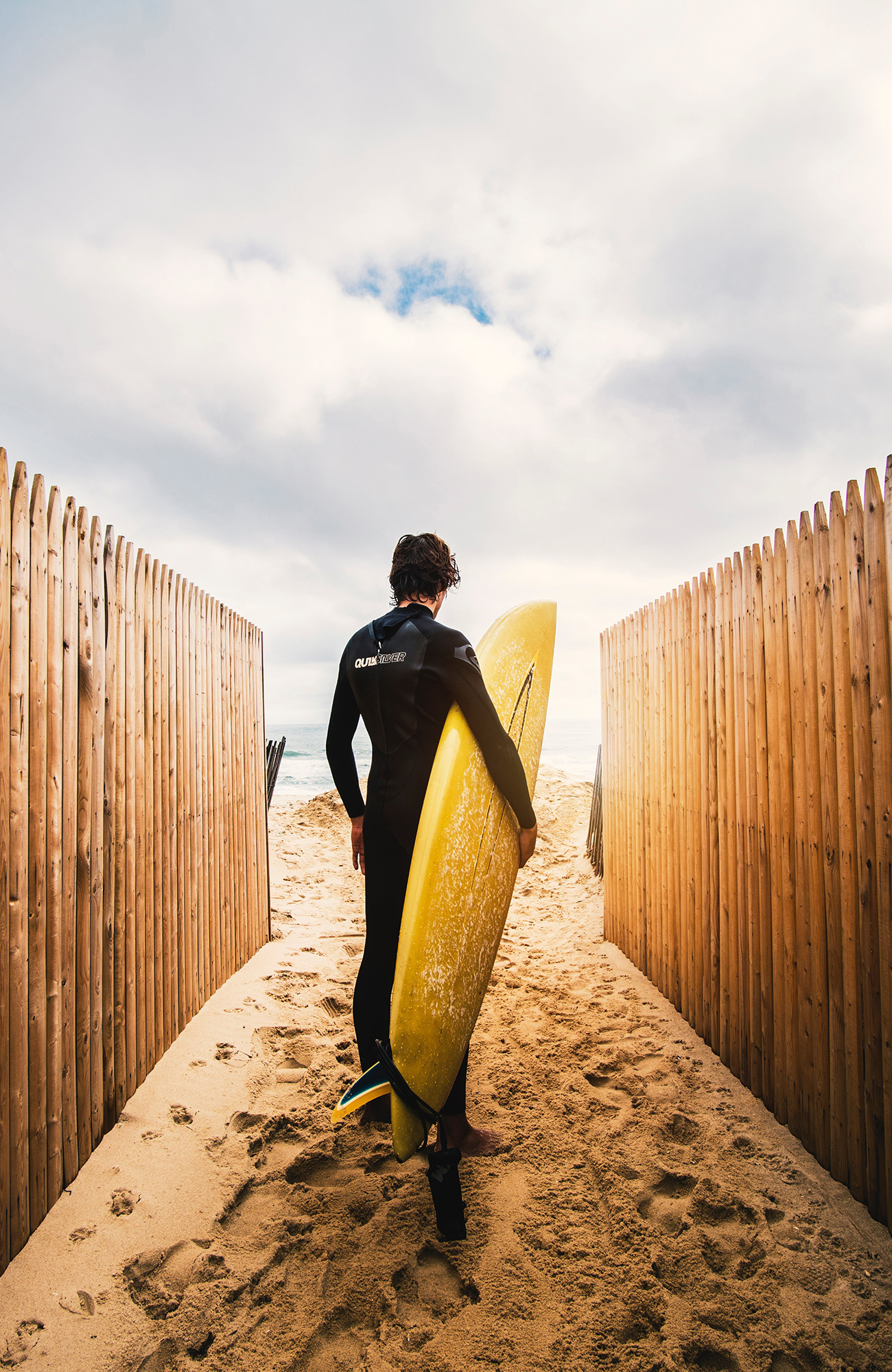 Image of a man in a wetsuit holding a yellow surfboard and standing in the sand - KILROY