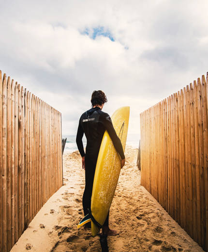 Image of a man in a wetsuit holding a yellow surfboard and standing in the sand - KILROY