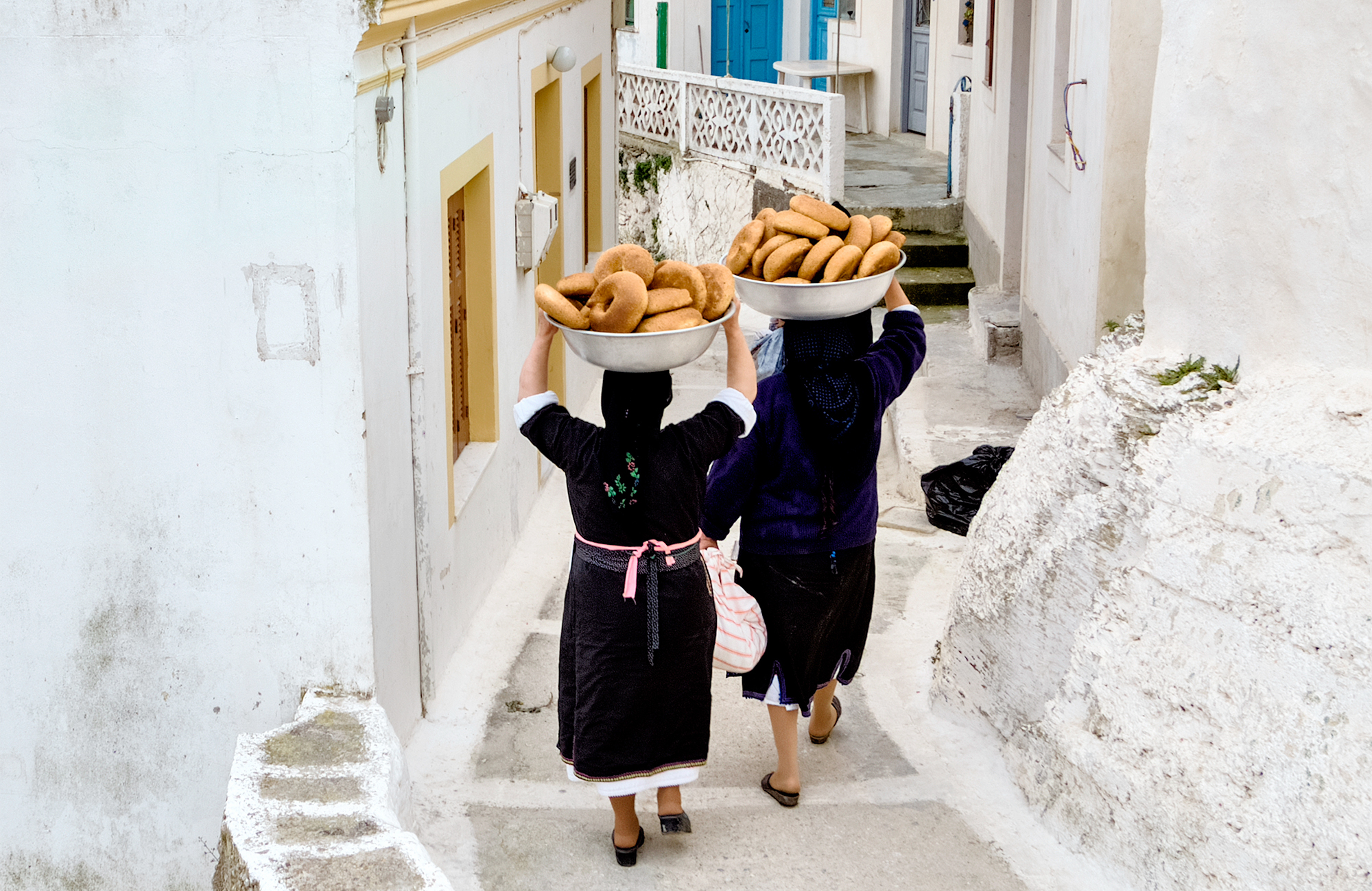 Image of local women carrying large bowls of bread on their head as they walk through traditional streets in Greece - KILROY