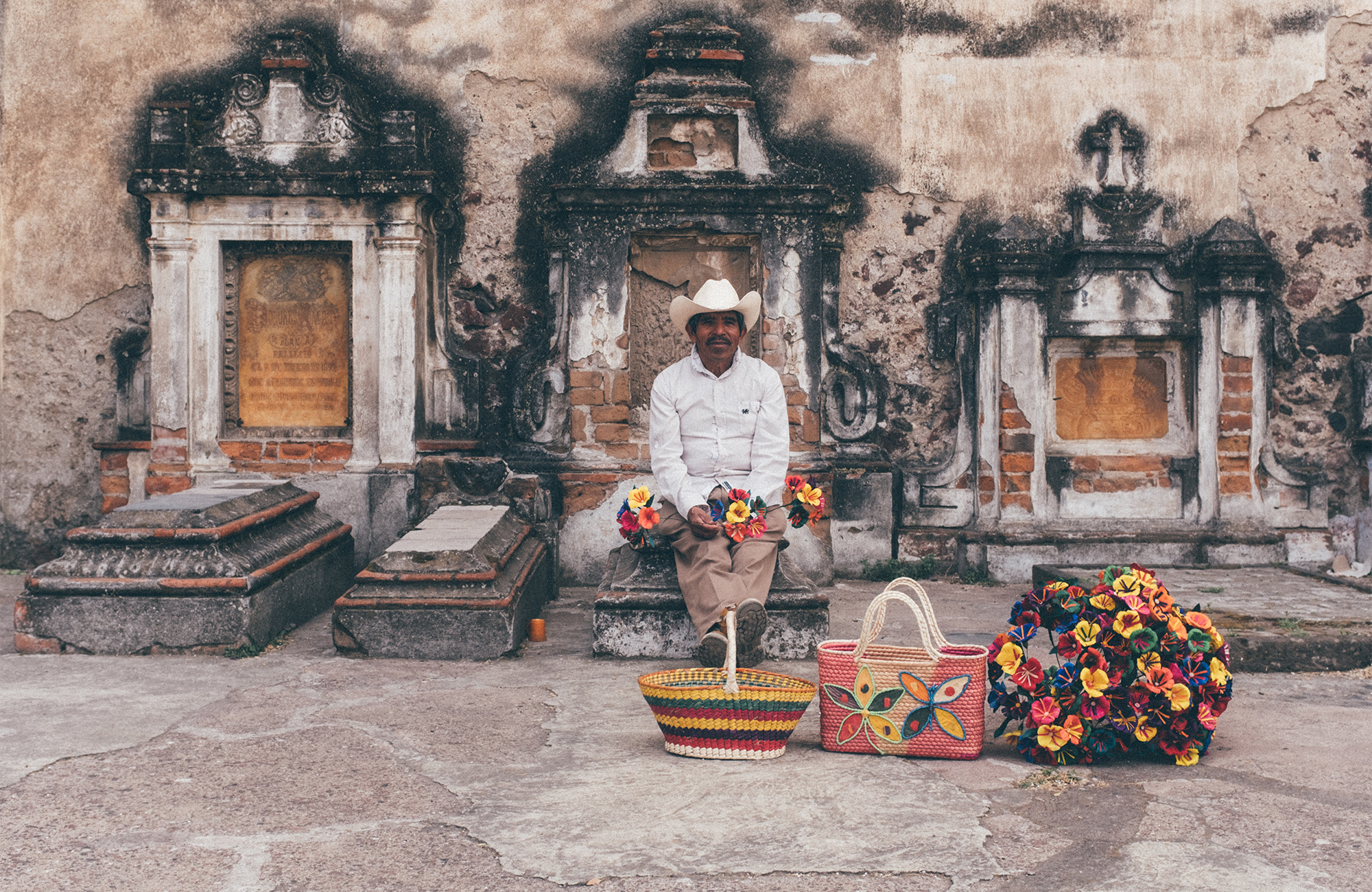 Image of a man sitting on a tomb with baskets of flowers in Mexico - KILROY
