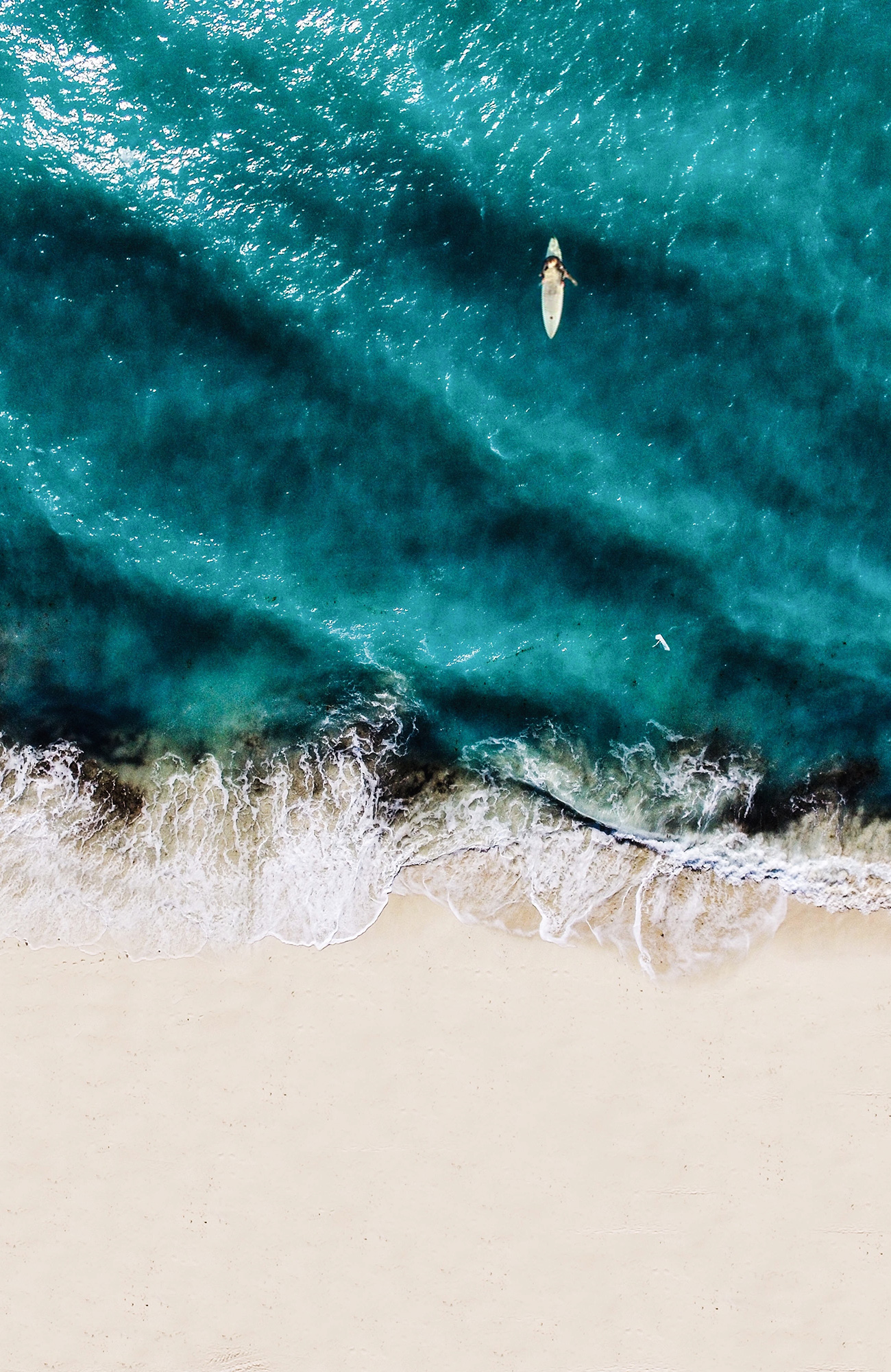 Aerial view of a surfer on the water as the waves break on a perfect sandy beach - KILROY