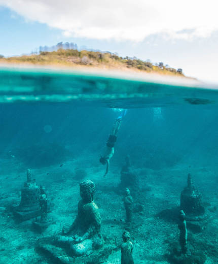 Image of a man diving under water where Buddhist statues decorate the sea bed - KILROY