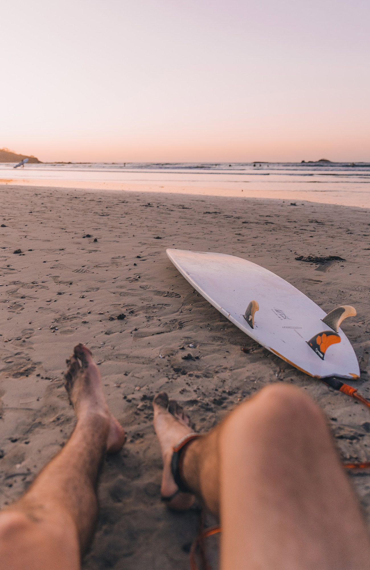 Image of a man's legs as he sites on the beach with a surfboard in front of him - KILROY