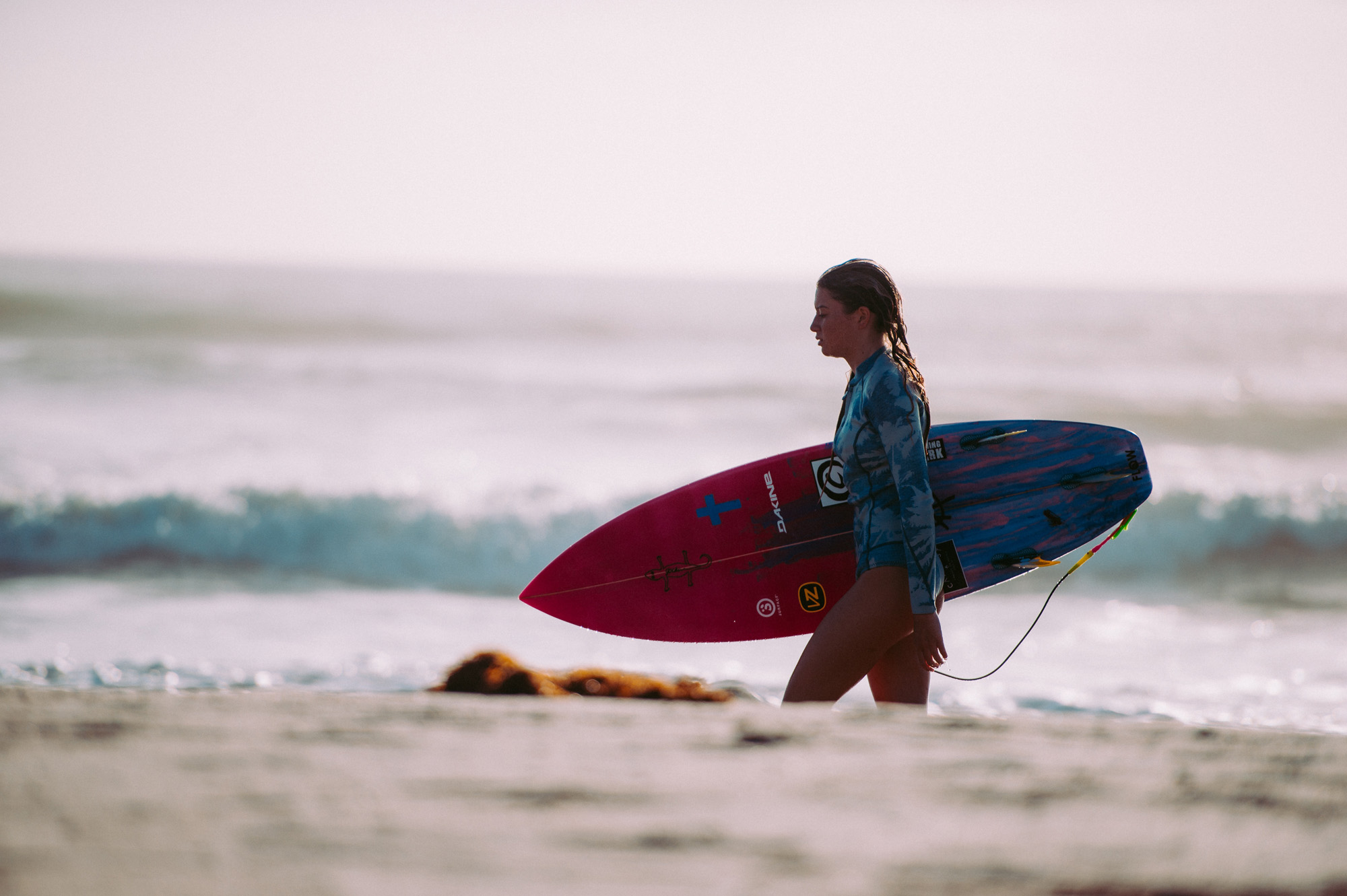 Image of a woman carrying a surfboard along a beach - KILROY