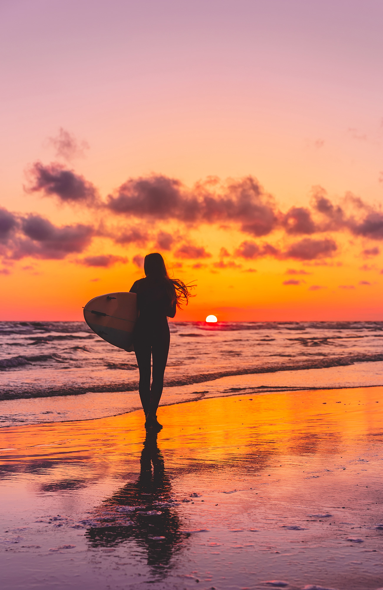Image of the silhouette of a woman carrying a surfboard on a beach at sunset - KILROY