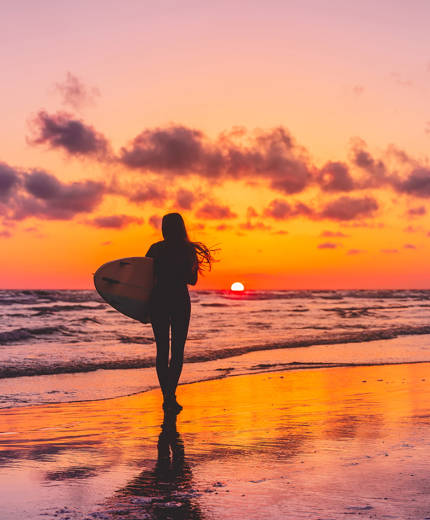 Image of the silhouette of a woman carrying a surfboard on a beach at sunset - KILROY