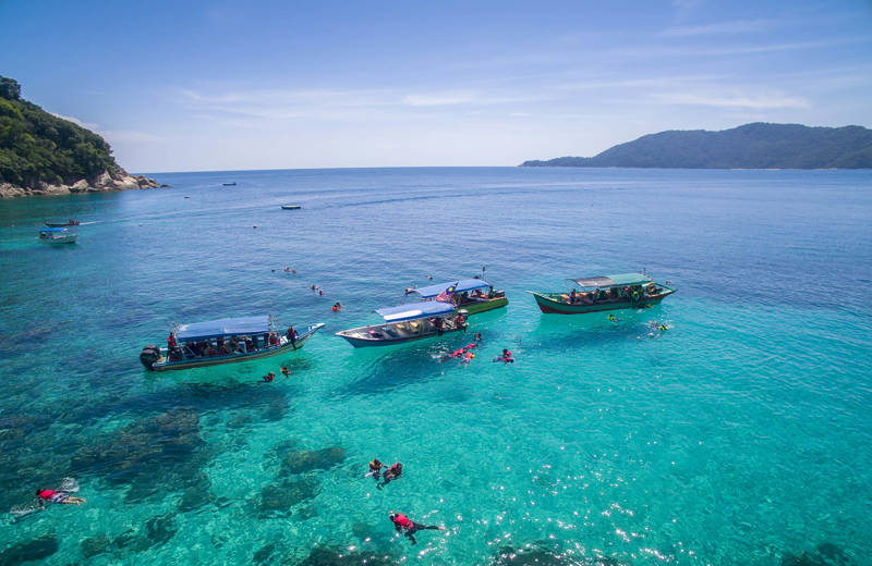 A couple of boats off the coast of the Perhentian islands, with people diving and snorkeling in the light blue water surrounding the boats