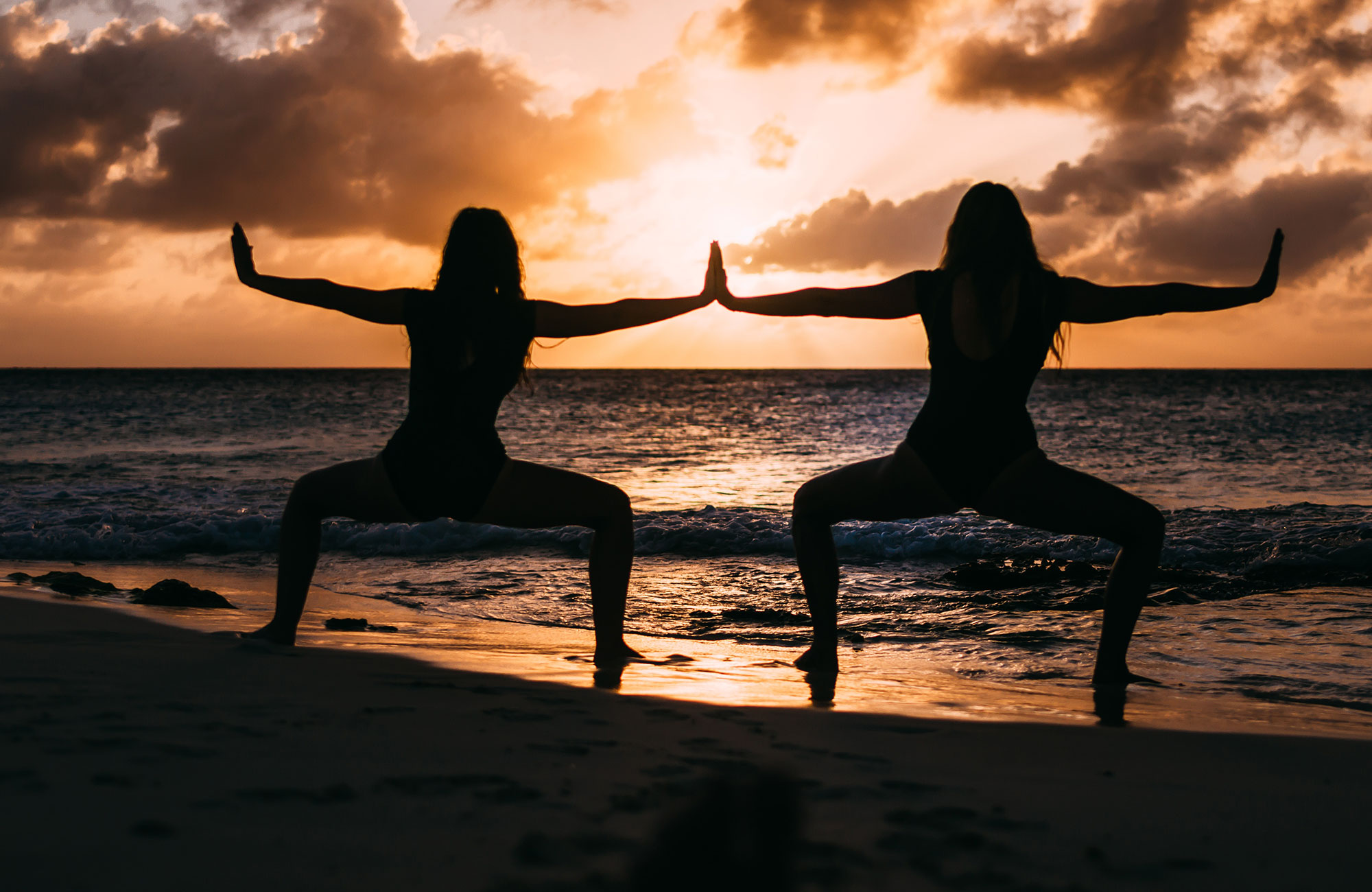 Image of the silhouette of two women holding a yoga pose at sunset on a beach - KILROY