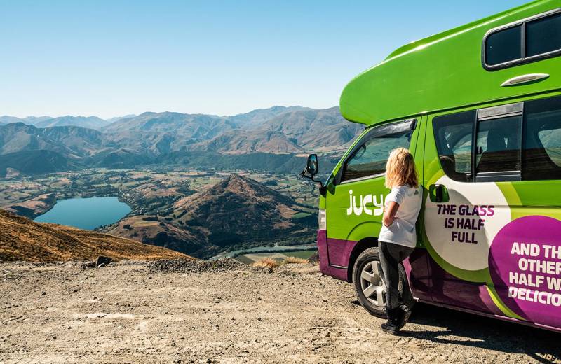 Girl leaning against a Jucy Condo Campervan, looking out over a lake and mountains