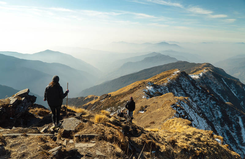 Two travellers hiking on a rocky mountain, with one traveller holding a stick and the other one looking at the mountainous view in the distance.