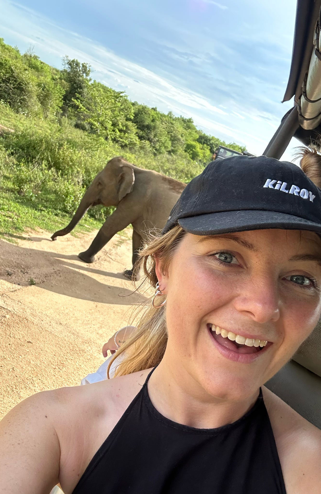 Image of a young woman wearing a KILROY cap in a safari truck with an elephant in the background - KILROY