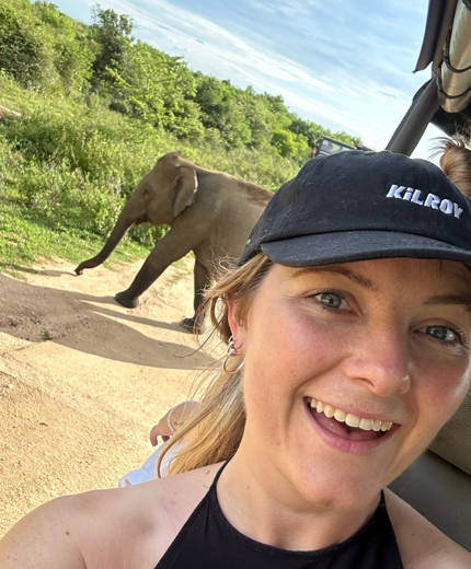 Image of a young woman wearing a KILROY cap in a safari truck with an elephant in the background - KILROY