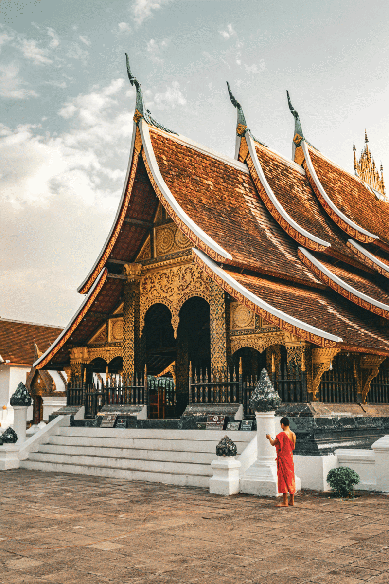 Monk dressed in an orange tunic standing in front of a temple in Luang Prabang in Laos