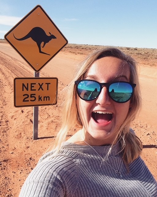 Image of a young female traveller next to a road sign in the Australian outback - KILROY