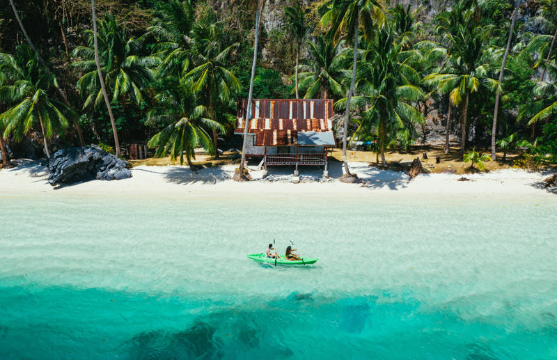 Image of a couple in a kayak alongside a palm-fringed beach in the Philippines - KILROY