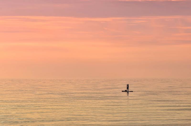 Man on a paddle board on a calm sea at dusk with a pink and purple sky - KILROY
